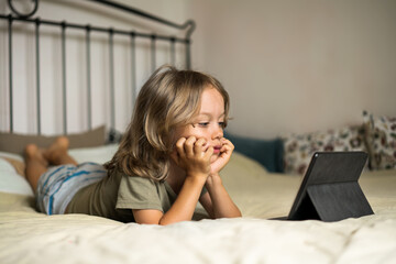 Boy lying on the bed using digital tablet computer playing games or watching cartoons at home. Child internet safety concept. Kid using electronic equipment. Parental control. Online education.