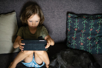 Boy sitting on sofa using digital tablet computer playing games or watching cartoons at home. Child internet safety concept. Kid using electronic equipment. Parental control. Online education.