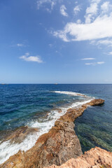 Vertical shot: narrow strip of rock gently descends into sea waters and is lost somewhere in distance, leaving foam from sea waves, Ibiza, Balearic Islands, Spain