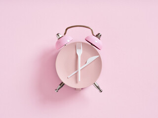 Alarm clock with plate and cutlery on pink background. Intermittent fasting, lunchtime, weight loss, meal plan or diet