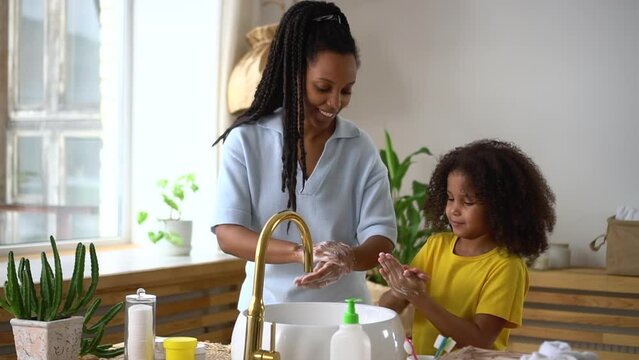 Young mother is teaching daughter to wash hands and smiling while standing in bathroom spbd. Close view of american african woman, cute girl clean arms and use soap, talk with smiles and stand by sink