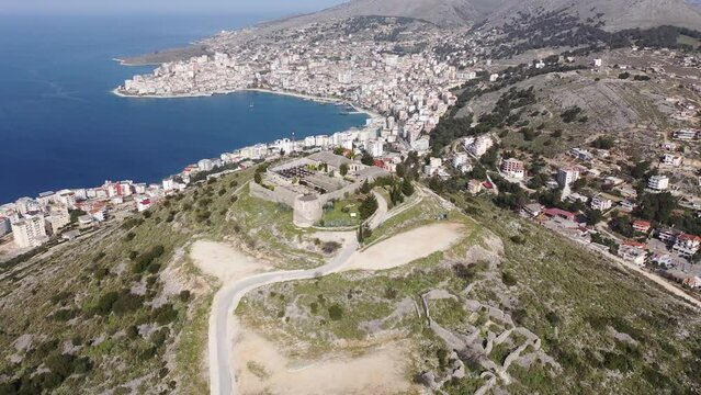 Scenic aerial view of Sarande cityscape on shores of gulf of Ionian Sea and ancient Lekuresi Castle on hill above city on sunny spring day, Albania