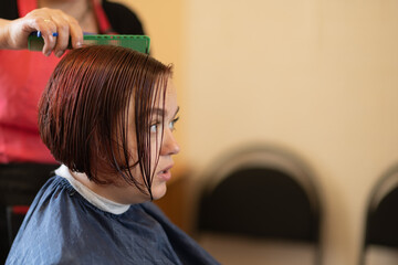 Naklejka premium A brown-haired girl is sitting in a chair at the hairdresser doing her hair and styling her hair.