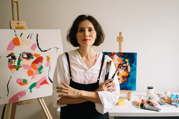Real Female Artist Dirty with Paint, Wearing Apron, Crosses Arms while Holding Brushes, Looks at...