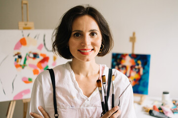 Real Female Artist Dirty with Paint, Wearing Apron, Crosses Arms while Holding Brushes, Looks at...