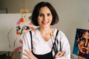 Real Female Artist Dirty with Paint, Wearing Apron, Crosses Arms while Holding Brushes, Looks at...