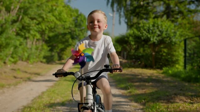 Cute child rides bicycle with a colorful windmill toy. Happy smiling blond caucasian boy rides along dirt road on white bicycle among green trees and spinning toy windmill on handlebars of bicycle.