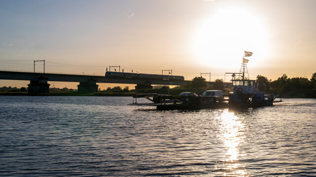 Silhouette of ferry boat, train and bridge early evening. Sunshine reflecting in the river. It is the ferry over the river Lek, at Culemborg, the Netherlands.
