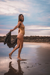 Fototapeta na wymiar A brunette woman having fun with a swimsuit and a pareo walking along the beach in summer in the cloudy sunset