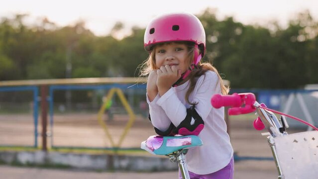 Portrait of yong beautiful caucasian light hair girl. She leans her elbow on the bike seat and looks into the camera. Kid wears pink helmet and elbow pads. Sport activity for children concept.