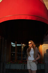 a woman in the sun at the building with red visors on the windows. city walks in summer, tourism. vacation in the city