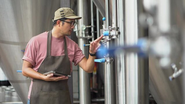 Asia male brewery worker with digital tablet in hands setting parameters on storage tank producing beer at factory.