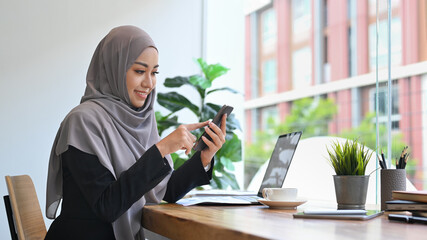 Smiling Muslim lady texting on mobile phone while working on computer laptop in office