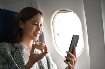 Beautiful asian businesswoman on a flight, eats a yummy doughnut and uses smartphone.