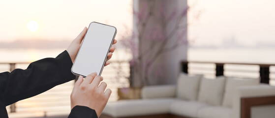 A female hand holding a smartphone mockup over blurred modern hotel balcony lounge