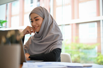 Cheerful young muslim woman wearing hijab at her office desk, looking at laptop screen