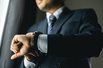 Confident businessman in classic blue suit wearing or adjust watch with necktie near window in...