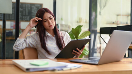 Serious young female employee sitting front of laptop and reading financial document