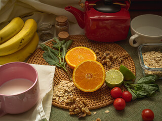 Healthy food still life with oranges, bananas, limes, milk, cereals, nuts, teapot, tablecloth and various cups lit with soft natural light