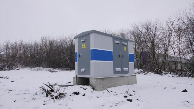 Small electrical transformer building with metal switchboards cabinets on snowy ground under sky with grey clouds