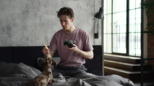 4k Young man is training dog and taking photo while sitting on bed at home spbd. Close view of caucasian guy trains cute pet and holds camera in hands, takes picture and sits in light interior