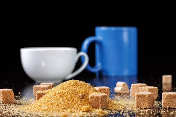 Brown cane sugar and pieces of sugar on the background of a cup and mug. Selective focus.