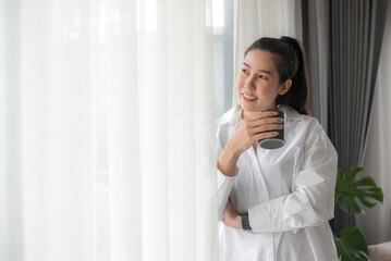 Smiling happy cheerful beautiful asian young woman relaxing and drinking hot coffee or tea and looking out of window. Girl felling enjoy having breakfast in holiday morning vacation in living room.