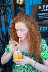 Young redheaded girl with frecked face holding fresh citrus summer ice cocktail
