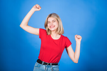 Young and attractive blonde caucasian girl in casual clothes dancing and celebrating victory, showing winner gesture isolated on blue studio background.