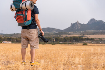 Hombre con mochila de acampada y esterilla, con su cámara de fotografía, en un paisaje de prados y montañas. Fotografía horizontal con espacio para texto.