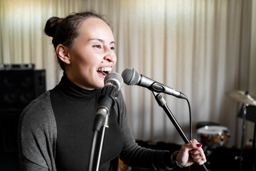 Young Japanese or Caucasian brunette woman having fun fooling around singing rock song on two microphones emotionally loud and with pleasure alone in music studio or school, event or concert stage.