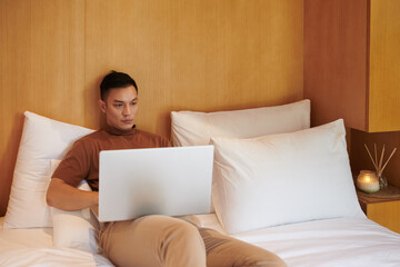 Asian young man working on laptop when sitting on comfy bed at home