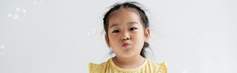 portrait of asian preschooler girl pouting lips near near soap bubbles isolated on grey, banner.