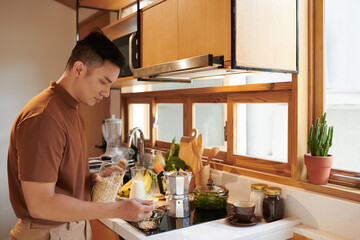 Asian young man adding two spoons on oatmeal in glass bowl when making breakfast