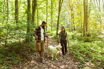 Obraz premium Young happy couple and their dog at hiking through the woods enjoying the sight. Two nature lovers in the mountain forest enjoy healthy walking through the nature. With film grain