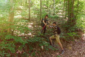 Young man helping his woman to step down on the steep hill in the forest while they hiking through...