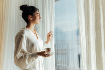 Attractive woman is holding a cup with hot tea or coffee and looking at the sunrise while standing...