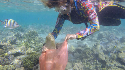 Woman in diving equipment swims and collects plastic debris underwater on the bottom of coral reef....