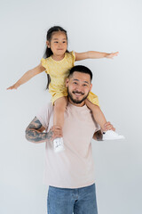 happy asian child with outstretched hands sitting on shoulders of tattooed father isolated on grey.