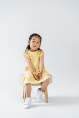 full length of asian kid in yellow dress and white sneakers sitting on chair on grey.