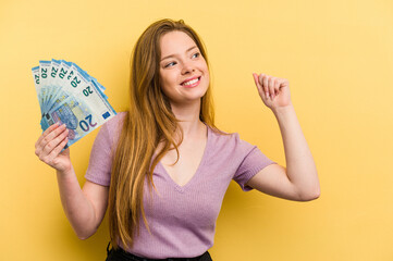 Young caucasian woman holding banknotes isolated on yellow background raising fist after a victory, winner concept.