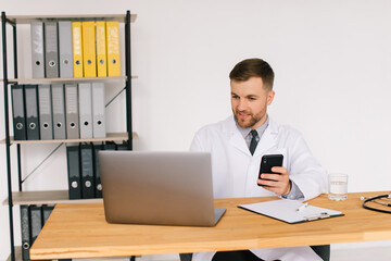 Male doctor holds a phone and sits in the office near the laptop