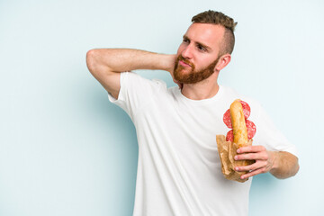 Young caucasian man eating a sandwich isolated on blue background touching back of head, thinking and making a choice.