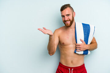 Young caucasian man going to the beach holding a towel isolated on blue background showing a copy space on a palm and holding another hand on waist.