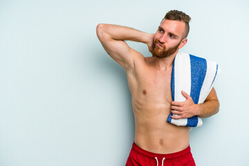 Young caucasian man going to the beach holding a towel isolated on blue background touching back of head, thinking and making a choice.