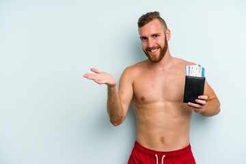 Fototapeta premium Young caucasian man holding a passport isolated on blue background showing a copy space on a palm and holding another hand on waist.