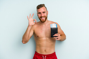 Fototapeta premium Young caucasian man holding a passport isolated on blue background cheerful and confident showing ok gesture.