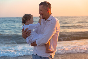 A young dad with a little daughter walks along the sea beach in the sunset.