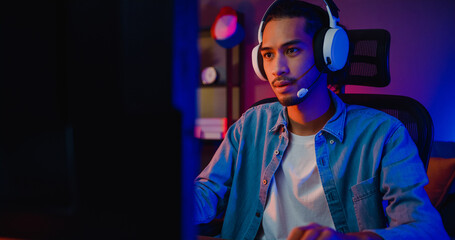 Happy asia guy gamer wear headphone participation play video game colorful neon lights computer in living room at night modern house. Esport streaming game online, Home quarantine activity concept. - obrazy, fototapety, plakaty
