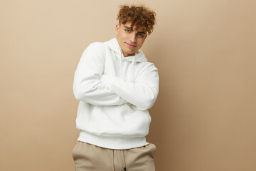 a stylish young man in a white hoodie stands on a beige background and pleasantly looking at the camera keeps his arms crossed on his chest